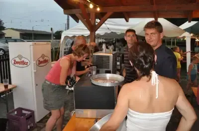 Eine Gruppe von Menschen steht um einen Imbisswagen draußen. Eine Frau in einem rosa Shirt lehnt sich über den Tresen und ein Mann in einem blauen Shirt steht hinter ihr. Andere Leute sind in der Nähe und ein Kühlschrank mit dem Wort Zwietller ist sichtbar.