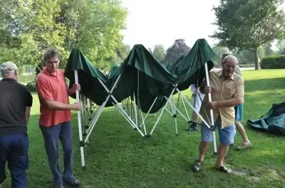 Mehrere Personen bauen ein grünes Zelt in einem Graspark mit Bäumen im Hintergrund auf. Sie tragen Freizeitkleidung.