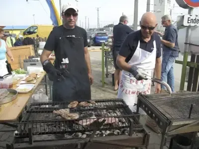 Zwei Männer grillen im Freien, einer benutzt Zangen. Hinter ihnen stehen mehrere Personen, einige tragen Hüte und Brillen. Im Hintergrund befindet sich ein Tisch mit Essen und ein Schild mit der Aufschrift 'Ende'.