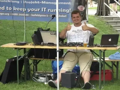 Ein Mann sitzt hinter einem Schreibtisch mit mehreren elektronischen Geräten und zeigt Daumen hoch. Der Schreibtisch hat zwei Laptops, Lautsprecher und einen Monitor. Im Hintergrund befindet sich ein Banner mit Text und Logos.