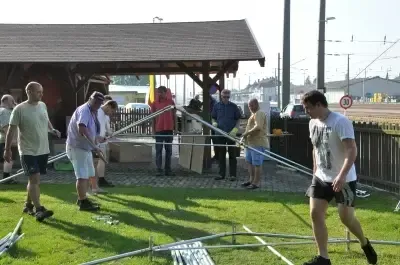 Eine Gruppe von Menschen baut eine Struktur auf einem Rasenfeld zusammen, mit einem Holzhaus im Hintergrund. Einige tragen Handschuhe und Turnschuhe. Der Himmel ist klar.