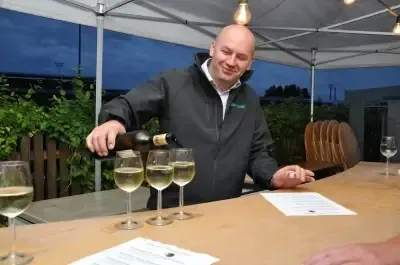 Ein Mann schenkt weißen Wein in Gläser auf einer Bartheke unter einem Zelt bei Dämmerung, umgeben von Grün und einem Zaun.