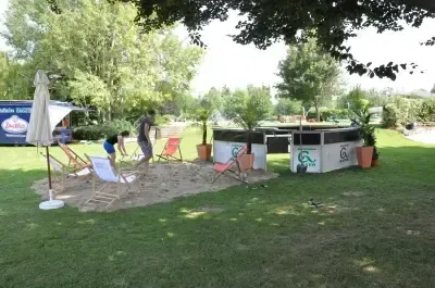 Ein Außenbereich mit einem sandigen Spielbereich, Liegestühlen und einem geschlossenen Schirm. Eine Person steht auf dem Sand und eine andere Person steht neben ihm. Es gibt Topfpflanzen und Bäume im Bereich. Ein blaues Schild ist an der Wand befestigt.