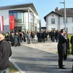 Eine Menschenmenge versammelt sich vor einem modernen Gebäude. Einige gehen, andere stehen. Es sind Flaggen und Banner ausgestellt. Das Gebäude hat große Glasfenster und ein modernes Design.