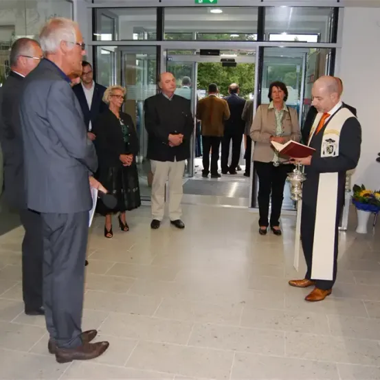 Eine formelle Veranstaltung mit Teilnehmern in Geschäftskleidung. Ein Mann in einem weißen Weste hält ein Buch, möglicherweise ein Priester, während ein anderer Mann ein Mikrofon hält. Sie stehen vor einer Glastür mit offenem Eingang.