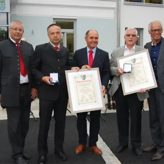 Fünf Männer stehen lächelnd vor einem Gebäude, mit Auszeichnungen und Urkunden. Sie tragen formelle Anzüge, einer mit roter Krawatte. Sie posieren für ein Foto.