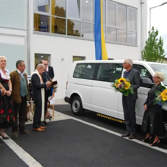 Eine Gruppe von Menschen steht um einen weißen Van, möglicherweise bei einer Enthüllungsveranstaltung. Einige halten Blumen, und ein Mann liest ein Buch. Hinter ihnen ist ein weißes Gebäude mit Glasfenstern zu sehen.