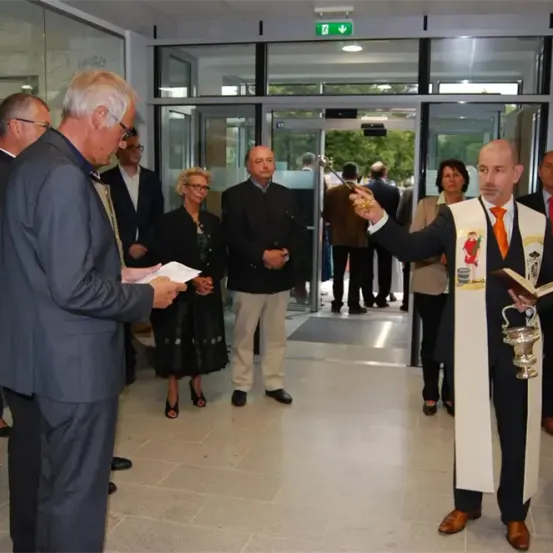 Eine Gruppe von Menschen in formeller Kleidung steht in einem Flur. Ein Mann in einer Robe hält ein Buch und einen Kelch. Ein älterer Mann überreicht einem jüngeren Mann ein Dokument. Hinter ihnen gehen Menschen durch eine Tür mit Glastüren in ein Gebäude.