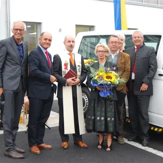 Mehrere formell gekleidete Personen stehen neben einem weißen Van, wobei eine Person einen Blumenstrauß und eine andere eine Buch hält.