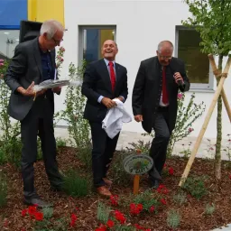Drei Männer in formeller Kleidung stehen in einem Garten. Einer hält einen Blumenstrauß, ein anderer ein weißes Tuch, und der dritte spricht in ein Mikrofon. Ein Baum und ein Schild sind in der Nähe.