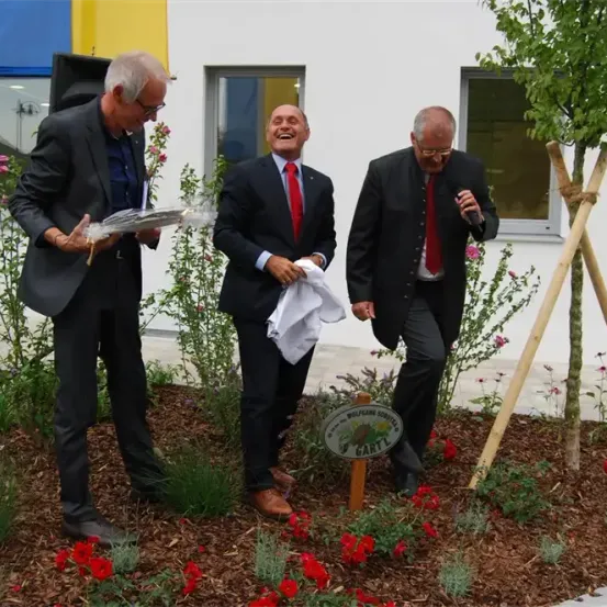 Drei Männer in formeller Kleidung stehen in einem Garten. Einer hält einen Blumenstrauß, ein anderer ein weißes Tuch, und der dritte spricht in ein Mikrofon. Ein Baum und ein Schild sind in der Nähe.