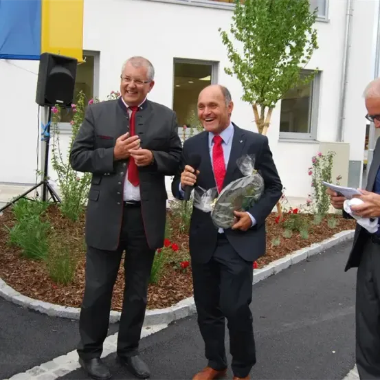 Drei Männer in Anzügen stehen vor einem Gebäude, einer hält ein Mikrofon und ein anderer ein eingewickeltes Geschenk. Sie scheinen bei einer Veranstaltung zu sein.