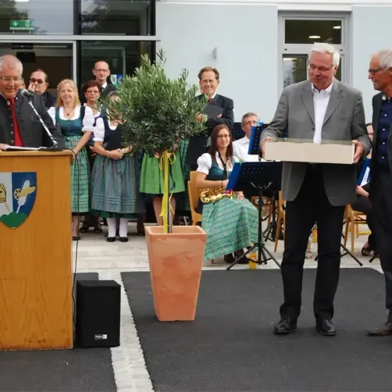 Ein Mann mit einem Mikrofon steht an einem Podium mit einem Wappen, während ein anderer Mann eine Schachtel hält und sie von einem dritten Mann erhält. Dahinter sitzt eine Frau auf einem Stuhl mit einem Saxophon.