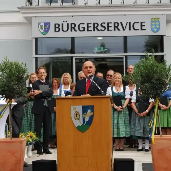 Ein Mann spricht an einem Podium vor einem Gebäude mit dem Schild Burgerservice. Hinter ihm stehen mehrere Personen.