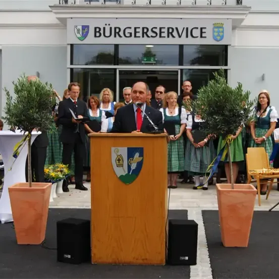 Ein Mann in Anzug und Krawatte spricht an einem Podium, mit einem Mikrofon vor ihm. Topfpflanzen und Banner befinden sich auf beiden Seiten. Eine Menschenmenge steht hinter ihm und das Gebäude trägt die Aufschrift Burgerservice.
