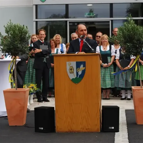 Ein Mann spricht an einem Podium mit Mikrofon. Hinter ihm stehen zwei Bäume in Töpfen. Ein Publikum beobachtet, gekleidet in traditioneller Kleidung.
