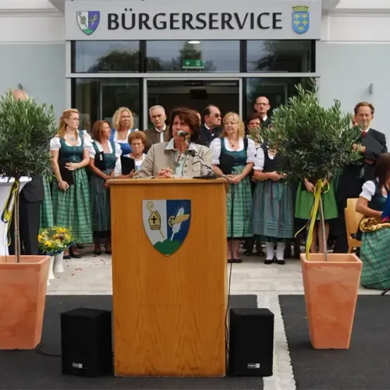 Eine Frau spricht an einem Podium vor einem Gebäude mit der Aufschrift Burgerservice, mit einer Gruppe von Menschen in traditioneller deutscher Kleidung hinter ihr.