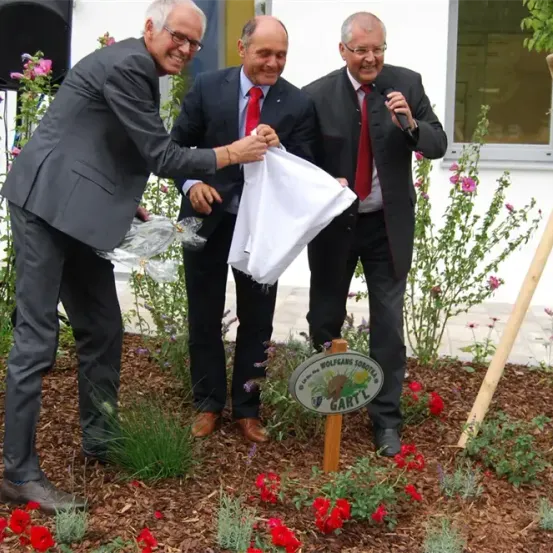Drei Männer in Anzügen und Krawatten pflanzen einen Baum, einer hält ein Mikrofon und ein anderer einen weißen Stoff.