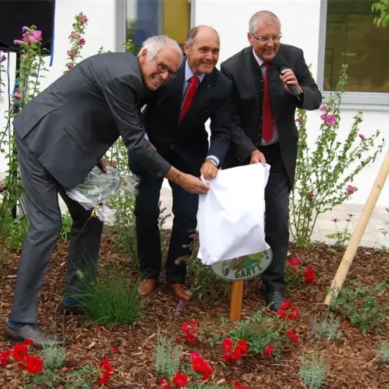 Drei Männer in Anzügen und Krawatten entfernen die Abdeckung eines kleinen Schildes in einem Garten.