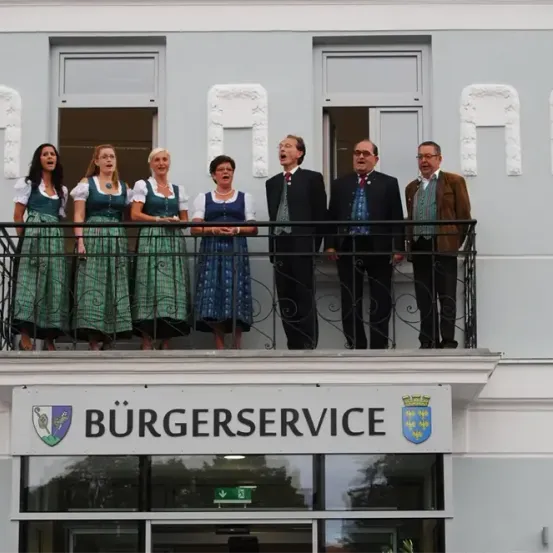 Eine Gruppe von Menschen, sowohl Männer als auch Frauen, steht auf einem Balkon und singt. Der Balkon befindet sich über einem Gebäude mit einem Schild, das 'BURGERSERVICE' besagt. Die Frauen sind in traditioneller Kleidung gekleidet. Die Männer tragen Anzüge und Krawatten. Hinter ihnen befinden sich Fenster mit kunstvollen Verzierungen.