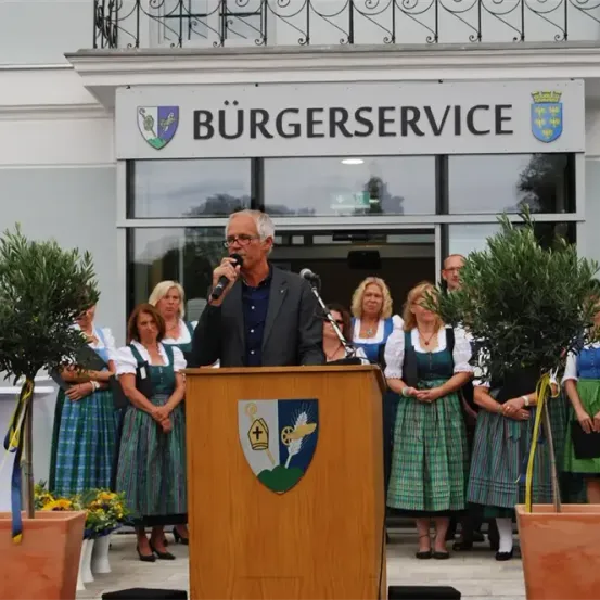 Ein Mann spricht an einem Podium vor einem Gebäude mit dem Schild Burgerservice. Hinter ihm stehen Frauen in traditioneller bayerischer Tracht und zwei Topfolivenbäume.