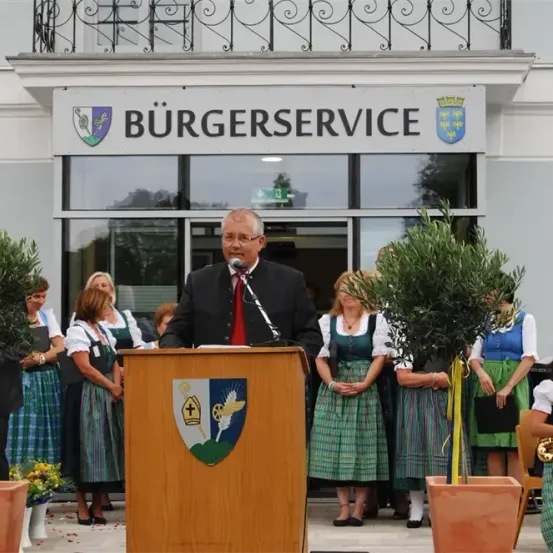 Ein Mann spricht an einem Podium vor dem Burgerservice-Gebäude, mit Frauen in traditioneller Kleidung hinter ihm. Topfpflanzen und Blumen schmücken den Bereich.