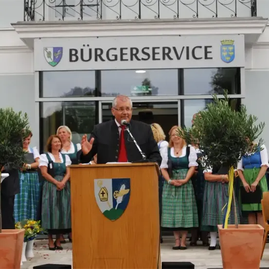 Ein Mann spricht an einem Podium mit einem Mikrofon, trägt einen Anzug und eine Krawatte. Dahinter steht ein Schild mit der Aufschrift Burgerservice. Im Hintergrund stehen Menschen in traditionellen grünen und weißen Kleidern, mit Bäumen und einer Balkon.