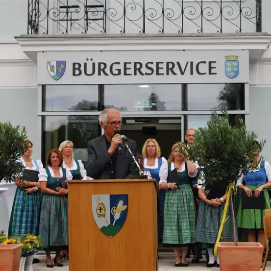 Ein Mann spricht an einem Rednerpult mit einem Mikrofon vor einem Gebäude mit dem Schild Burgerservice, mit mehreren Frauen in grünen und weißen Kleidern hinter ihm.