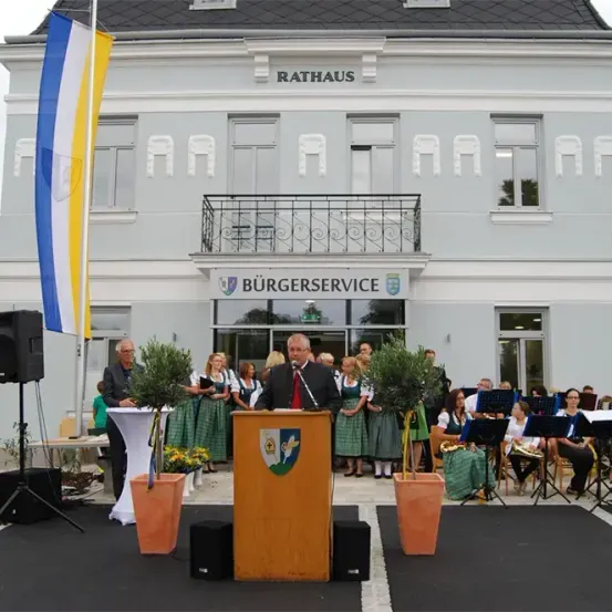 Ein Mann hält eine Rede an einem Rednerpult vor einem Gebäude. Das Gebäude hat ein Schild mit der Aufschrift Rathaus Burgerservice. Hinter dem Rednerpult sitzen Menschen auf Stühlen mit Musikinstrumenten.