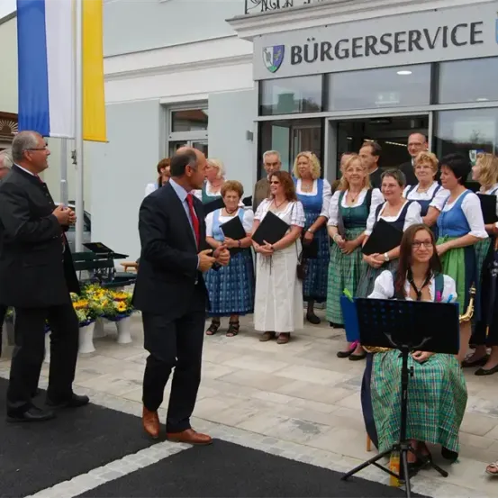 Eine Gruppe von Menschen steht vor einem Gebäude mit der Aufschrift Burgerservice. Ein Mann spricht mit einem anderen Mann. Eine Frau spielt Saxophon. Eine Frau sitzt auf einem Stuhl. Es gibt Blumen auf dem Boden. Es gibt zwei Fahnen an Stangen.