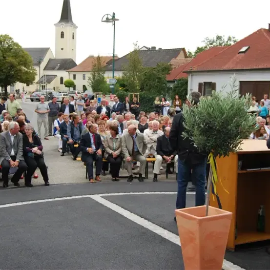 Eine Menschenmenge sitzt auf Bänken und hört einem Redner an einem Podium zu. Eine Kirche und Häuser sind im Hintergrund.