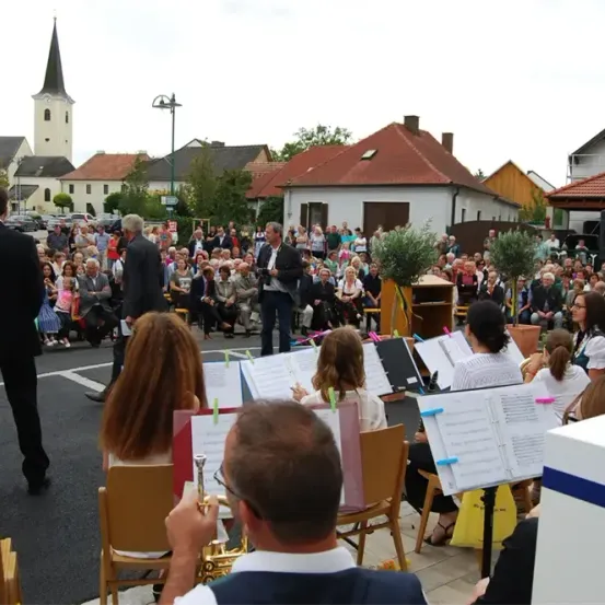 Eine Gruppe von Musikern tritt in einer Freiluftumgebung auf, mit einem großen Publikum, das auf Stühlen sitzt. Ein Dirigent leitet, und dahinter ist ein Gebäude mit einem Kirchturm zu sehen. Die Umgebung umfasst Bäume, geparkte Autos und eine Straßenlaterne.