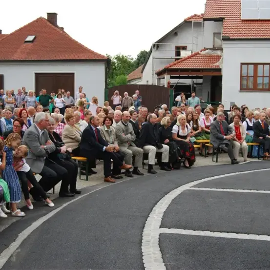 Eine Menschenmenge sitzt auf Bänken draußen, mit Häusern und Solarpanelen im Hintergrund.