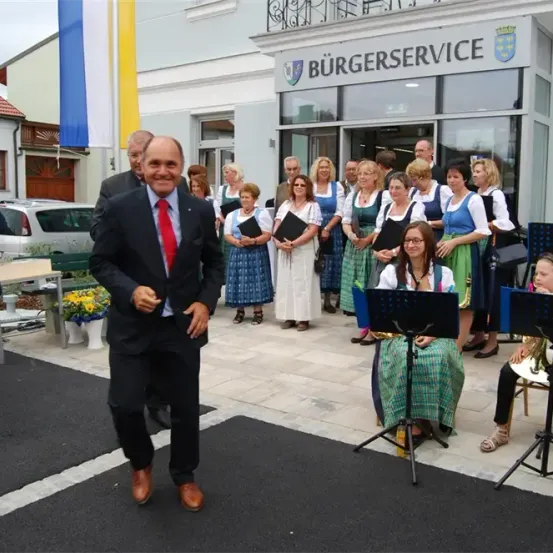 Eine Gruppe von Menschen in traditioneller Kleidung steht vor einem Gebäude namens Burgerservice. Ein Mann in Anzug geht auf sie zu. Musikinstrumente sind aufgestellt, und es gibt Pflanzen und ein Auto im Hintergrund.