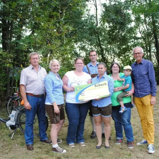 Eine Gruppe von Personen steht im Freien, ein Mann hält einen großen Scheck und im Hintergrund befinden sich Fahrräder.