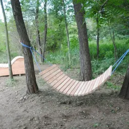 Ein hölzernes Hängemattenbett ist zwischen zwei Bäumen in einem Wald aufgehängt. In der Nähe befindet sich auch eine Holzbank.