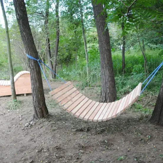 Ein hölzernes Hängemattenbett ist zwischen zwei Bäumen in einem Wald aufgehängt. In der Nähe befindet sich auch eine Holzbank.