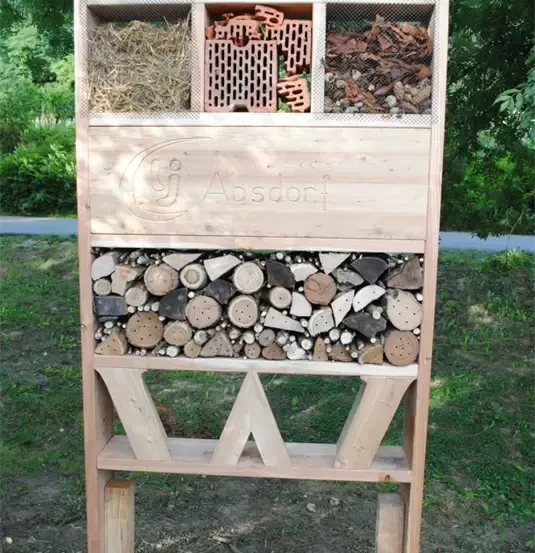 Ein hölzerne Außenlagereinheit mit drei Abschnitten. Der oberste Abschnitt hat Heu, der mittlere hat Ziegelsteine und der unterste hat Baumstämme. Sie steht auf einem Rasenfeld.