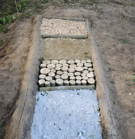 Ein hölzerner Weg mit geschichteten Materialien, darunter Kies, Holzspäne und eine Mischung aus pulverigen und steinigen Substanzen, in einem Erdfeld.