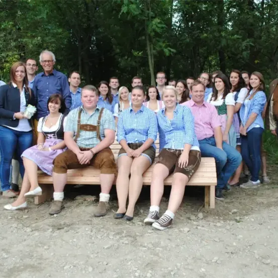 Eine Gruppe von Menschen in traditioneller bayerischer Kleidung posiert für ein Foto auf einer Holzbank im Wald.