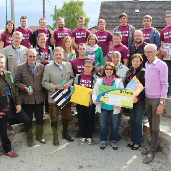Eine Gruppe von Menschen in passenden T-Shirts steht zusammen, einige halten Bücher und einen Scheck. Sie stehen vor einem Gebäude mit Bäumen im Hintergrund.