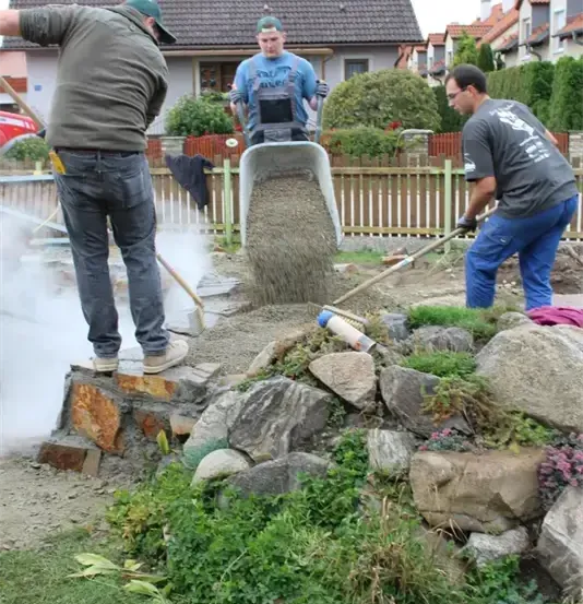 Drei Männer arbeiten auf einer Baustelle. Einer gießt Zement, ein anderer verteilt ihn und der dritte fegt. In der Nähe steht ein Haus und ein Zaun.