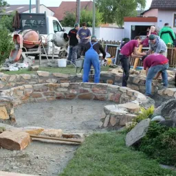 Eine Gruppe von Arbeitern baut eine Steinmauer in einem Hof. Zwei Männer benutzen eine Schubkarre. Zwei Zementmischer sind neben einem weißen Lieferwagen geparkt.