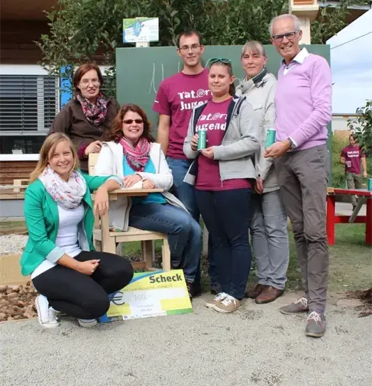 Eine Gruppe von Menschen posiert für ein Foto im Freien, in der Nähe eines Baumes und eines Gebäudes. Sie lächeln und halten Tassen.