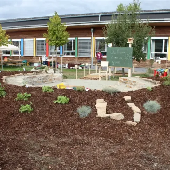 Ein Außenbereich mit einem Garten vor einem Gebäude mit mehreren Fenstern. Der Garten hat Pflanzen und Steine und eine Tafel steht in der Mitte.