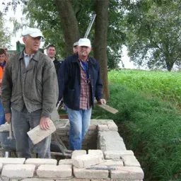 Fünf Männer bauen eine Ziegelmauer. Einer hält eine Kelle, und ein anderer raucht. Dahinter stehen zwei Bäume und ein grünes Feld.