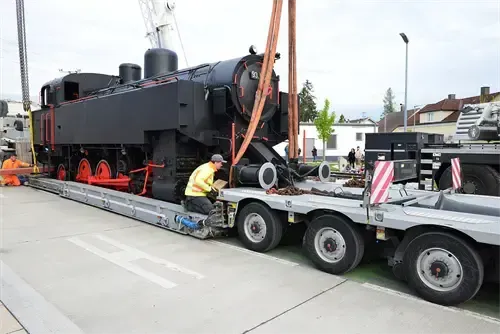 Eine schwarze Dampflokomotive wird an einem sonnigen Tag von einem Flachbett-LKW entladen.