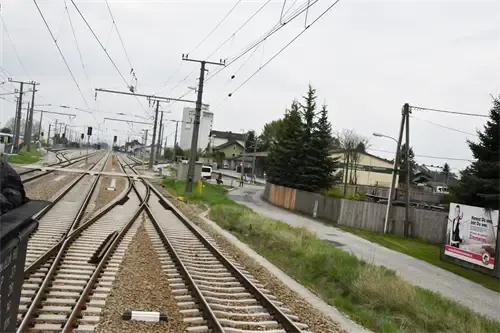 Eine Außenszene mit mehreren Bahngleisen, einem entfernten Gebäude, Bäumen und einer stehenden Person. Der Himmel ist bedeckt.