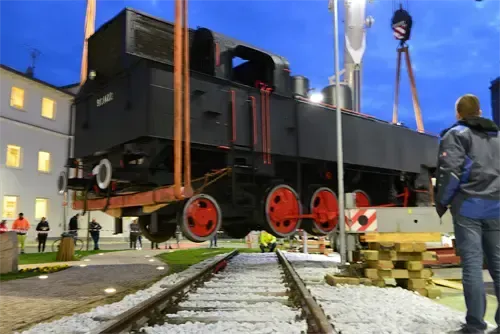 Eine Dampflokomotive wird von einem Kran auf eine Eisenbahnschiene gezogen. Zuschauer beobachten hinter einer Absperrung.