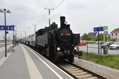 Eine alte Dampflokomotive mit der Nummer 93.1420 hält an einem Bahnhof. Der Bahnsteig hat Schilder mit der Aufschrift 'A 1 A'. In der Nähe sind Autos geparkt.